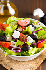 Fresh Greek salad made of cherry tomato, ruccola, arugula, feta, olives, cucumbers, onion and spices. Caesar salad in a white bowl on wooden background. Healthy organic diet food concept.