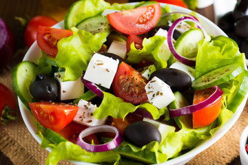 Fresh Greek salad made of cherry tomato, ruccola, arugula, feta, olives, cucumbers, onion and spices. Caesar salad in a white bowl on wooden background. Healthy organic diet food concept.