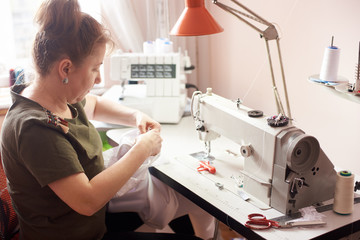 Clothier sitting in atelier, stitching white dress with hands and needle. Seamstress workplace with...