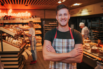 Young strong handsome man stand in grocery store or bakery. He crossed hands and smile. Positive worker. Young woman work behind close to bakery shelf.