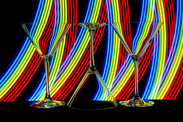Three cocktail / martini glasses in a row isolated against a black background with colorful streaks neon of light painting behind