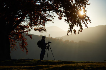 Profile of traveler tourist guy with backpack at camera tripod lit by bright setting sun in the sky standing on grassy valley on background of beautiful mountain panorama at dusk under big tree.