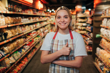 Happy positive young woman stand in line in grocery store. She look on camera and smile. Model keep big thumb up.