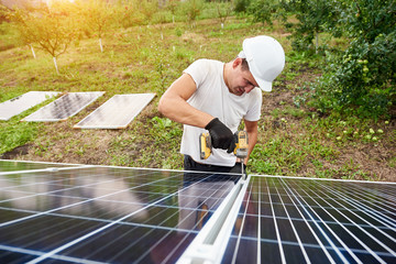 Young technician working with screwdriver connecting solar photo voltaic panel to exterior metal platform on sunny green orchard background. Alternative renewable sun energy sources concept.
