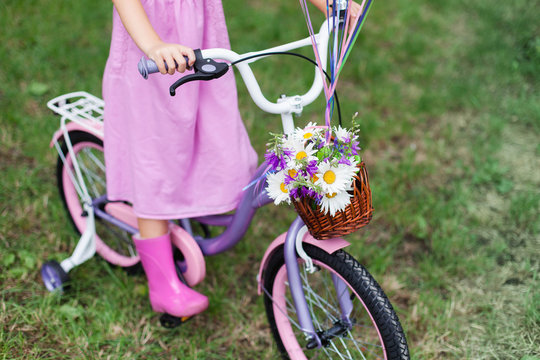 Little girl is riding on lilac bicycle with wicker basket with bouquet of summer flowers. Kid is wearing in pink dress and rubber boots. Child is playing in park.