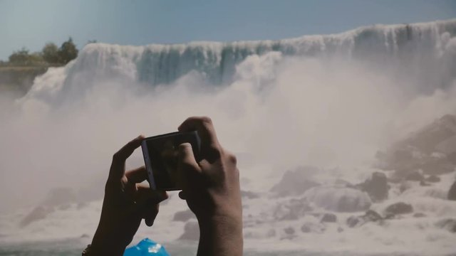 Slow motion close-up human hands taking smartphone photos of epic Niagara Falls waterfall panorama on a clear summer day