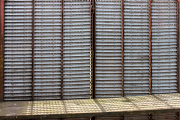 A fence of metal Selhozholding machinery parts, harvesters. The effect of blinds through which light passes. Horizontal details of gray color, divided into vertical sections. Next to the old wooden be