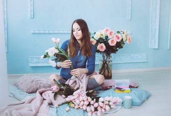 Cheerful young woman sitting on the floor, making flower bouquet of pink tulips in light sunlit room with blue walls.