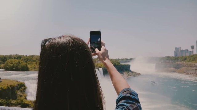 Slow motion camera slides left behind excited young tourist woman taking phone photo of amazing Niagara waterfall view.