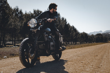 Young hipster boy riding a big classic cafe racer motorcycle in the forest with sunglasses