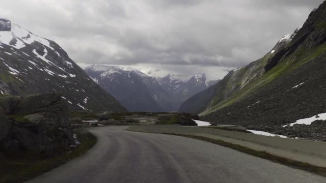 Driving down Gamle Strynefjellsvegen Riksvei 258 the old mountain road in Strynefjell, Oppland in the Norwegian mountains with snow and ice covered mountains in the background at a cloudy day.