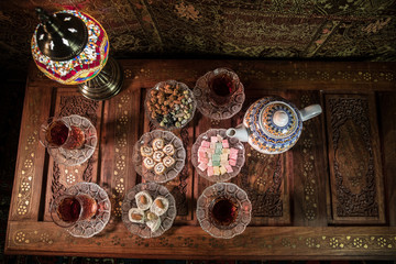 Arabian tea in glass with eastern snacks on vintage wooden surface. Eastern tea concept. Low light lounge interior with carpet. Empty space.