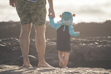 View from behind of legs of a young father with small child holding hands while looking at the sea...