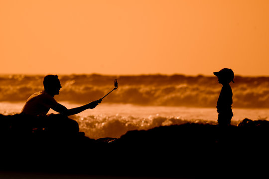 Silhouette father and son playing near the sea at the sunset. Concept of friendly family. Picture made on the background of orange sky. Family leisure time. Young male filming a child with smartphone.