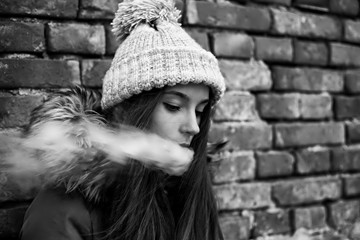 Vape teenager. Young pretty white girl in a cap and jacket smoking an electronic cigarette opposite brick wall on the street in the spring. Bad habit. Black and white.