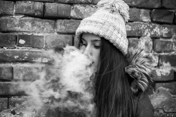 Vape teenager. Young pretty white girl in a cap and jacket smoking an electronic cigarette opposite...