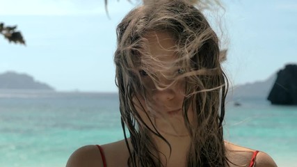 Close-up portrait of beautiful european young woman looking in the camera, hair blowing in wind on tropical beach, blue sea on the background. Travel concept.