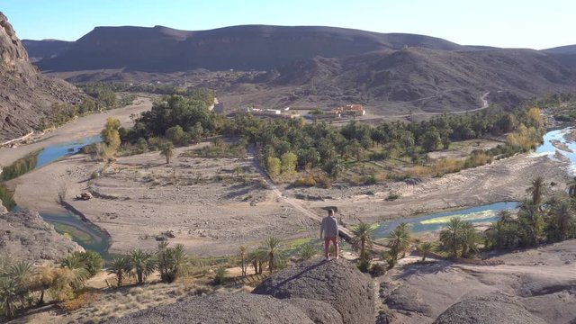 Traveller Guy Enjoying on High Rock Viewpoint at Beautiful Desert oasis landscape Panorama in Oasis De Fint near Ourzazate in Morocco, North Africa
