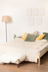 room with a light bed, a floor lamp and a photoframework on a wall. multi-colored pillows, yellow and green color on a bed