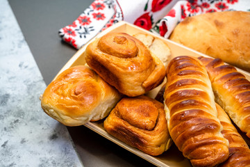 Tasty baked homemade buns with cinnamon and various fillings laid out on wooden plate and bread on towel with Ukrainian national ornament embroidery