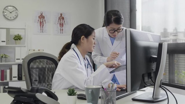 two doctors having informal meeting in hospital office. asian woman coworker walking close to ask for help on digital tablet solve problem while her colleague is typing on computer. girl nurses work