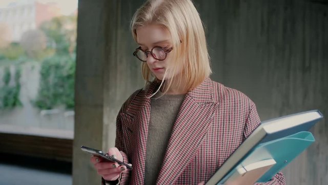 Portrait of young blond caucasian student girl in brown coat with textbooks checking publishing post on social network website or app reading digital book encyclopedia or news feed