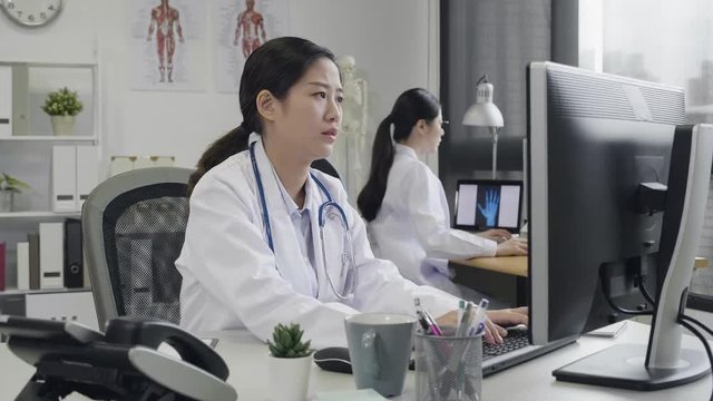 two medicine doctors writing typing on computer in medical office. asian female worker nurse wear stethoscope sitting at working desk in hospital concentrated using desktop pc. coworker next to her.