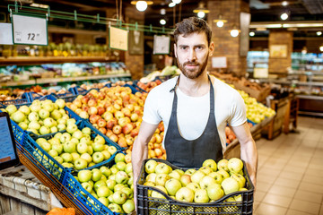 Portrait of a handsome worker or farmer in uniform holding box full of green apples in the shop or supermarket
