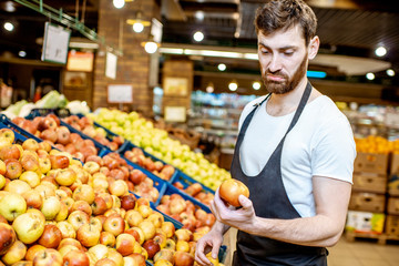 Handsome shop worker in uniform checking quality of the apples in the department with fruits and vegetabes in the supermarket