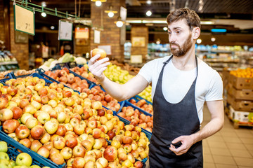 Handsome shop worker in uniform checking quality of the apples in the department with fruits and vegetabes in the supermarket