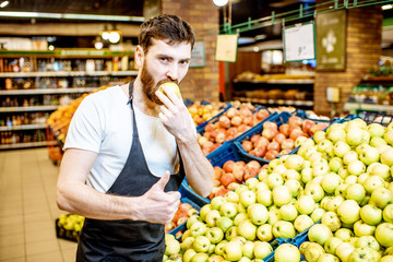 Handsome shop worker in uniform tasting apple checking the quality in the supermarket