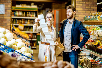 Young couple buying food standing together with long shopping list in the vegetable department of the supermarket. Man with shocked emotions - obrazy, fototapety, plakaty