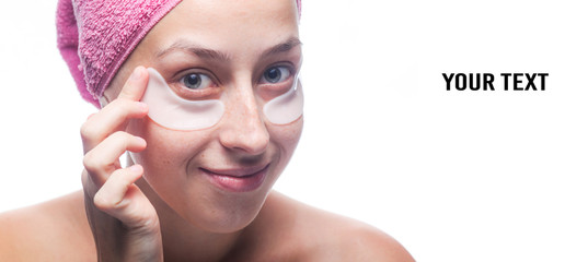 Copy space. Attractive smiling young woman with white patches under the eyes and a pink towel on her head isolated on white background. Closeup portrait. Skin care.
