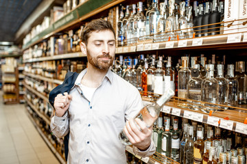 Portrait of a man with a thirst for alcohol, standing near the shelves with strong drinks in the supermarket