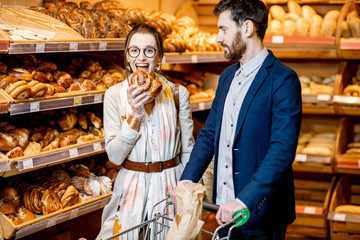 Young and happy couple choosing fresh pastries, standing together with shopping cart in the bakery department of the supermarket