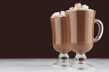 Christmas hot chocolate in two irish coffee glass with marshmallows and silver spoons on dark brown and white background. Hot holiday beverage.