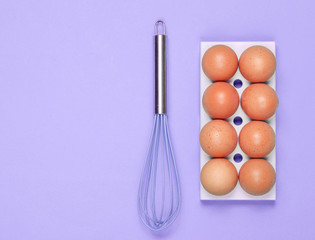 Silicone kitchen whisk, tray of chicken eggs on purple background. Cooking concept, Top view, flat lay, minimalism.