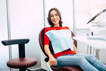Ready to roll. Thin cute girl clothed in red sweater and denim jeans is sitting in the dental chair...