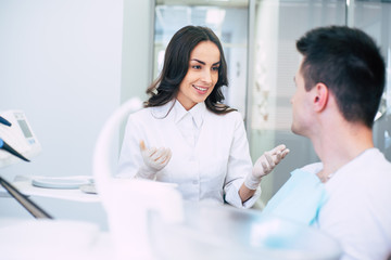 Fototapeta premium Dental clinic. Professional dentist in spacious dentist room is wearing medical clothes and giving some recommendations to her patient after his appointment.