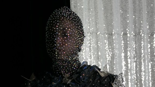 High-fashion. Model in the image of an alien girl in a black mask in sparkling rhinestones, sparkles, metal butterflies on a black and silver background posing in a model studio