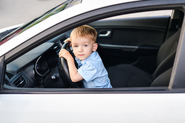 A blond boy with blue eyes sits in the front seat behind the wheel of a white car..