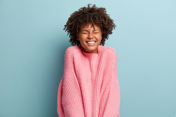 Joyful satisfied black woman with curly bushy haircut, expresses good emotions and feelings, has toothy smile, being under impression, laughs sincerely, wears oversized pink jumper, stands indoor