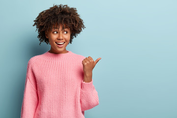 Indoor shot of lovely Afro woman points away with thumb, suggests using copy space wisely, wears...