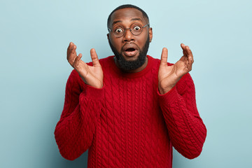 Stupefied black young man gestures with hesitation, has negative reaction on something horrified,...