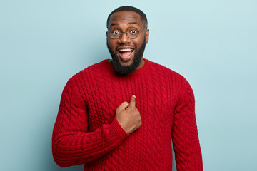 Really me are you serious? Cheerful dark skinned entrepreneur being picked for promotion, points at chest with index finger, asks question, isolated over blue background. Male indicates at himself