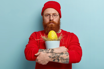 Indoor shot of handsome man with thick foxy bristle, carries tightly colored egg in hay, has tatooed arm, looks seriously at camera, wears red headgear and jumper, models over blue background