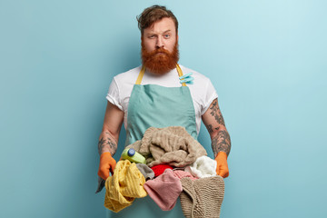 Laundry day concept. Serious bearded red haired Caucasian man busy doing housework, holds basket...