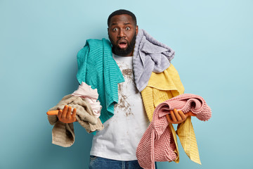 Isolated shot of indignant black ethnic man has bristle overloaded with laundry, stares with bugged...