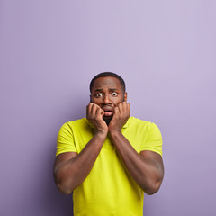 Omg, my time is up. Nervous dark skinned guy keeps hands near mouth, has frustrated look, bites fingernails from puzzlement, expresses negative feelings, stands against purple wall with empty space