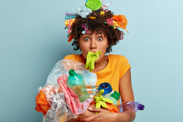 Taking care of environment. Emotive surprised dark skinned woman stares with bugged eyes, organizes local event for cleaning planet from garbage, keeps rubber glove in mouth, plastic things on hair